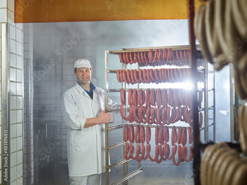 Smiling butcher standing by arranged sausages in smokehouse photo