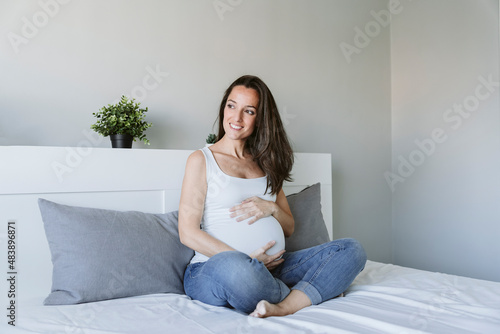 Happy pregnant woman with long brown hair sitting on bed at home
