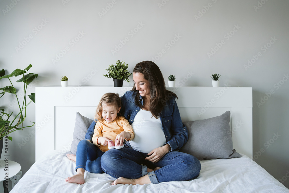 Happy pregnant woman looking at daughter holding baby booties on bed Happy pregnant woman looking at daughter holding baby booties on bed