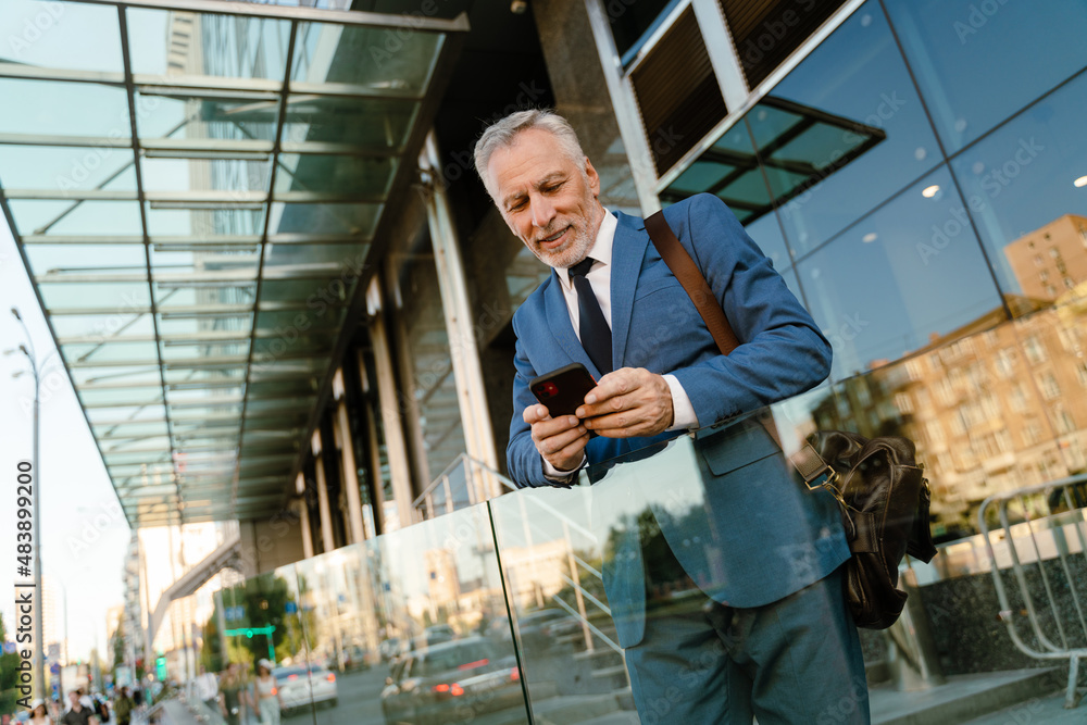 Senior man dressed in suit using mobile phone while standing by building