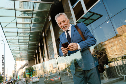 Senior man dressed in suit using mobile phone while standing by building