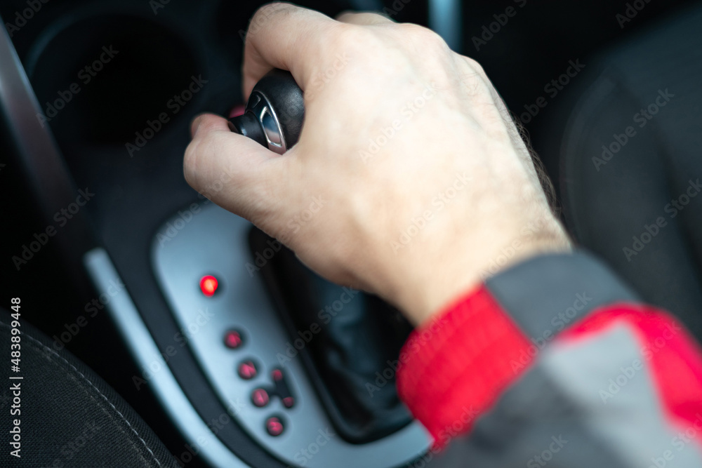 A man's hand in blurry focus is holding a gearbox in a parking lot.