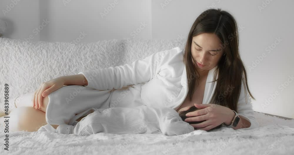 Caring mother is breastfeeding her newborn baby while lying on the sofa in the nursery. Mom in a white coat holds and cradles a infant in her arms. Concept of Childhood, New Llife, Parenthood. Caring mother is breastfeeding her newborn baby while lying on the sofa in the nursery. Mom in a white coat holds and cradles a infant in her arms. Concept of Childhood, New Llife, Parenthood.