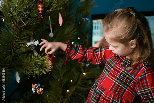 a girl in a red plaid dress hangs a nutcracker toy on the Christmas tree. Christmas Holiday Tree