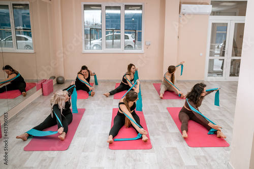 A group of six athletic women doing pilates or yoga on pink mats in front of a window in a beige loft studio interior. Teamwork, good mood and healthy lifestyle concept.