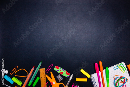 Series of stationery items, colored pens, labels, paper clips, calculator, scissors and more positioned below on black background, back to school concept, flat lay, blank space