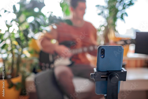 Young musician in cozy sweater sitting on the table and playing electric guitar live in front of the smartphone camera. Online concert during lockdown. Social distance learning. Focus on phone