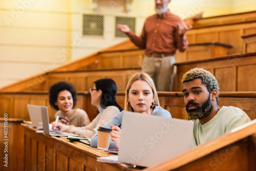 Multiethnic students looking at laptop near blurred friends and teacher in university