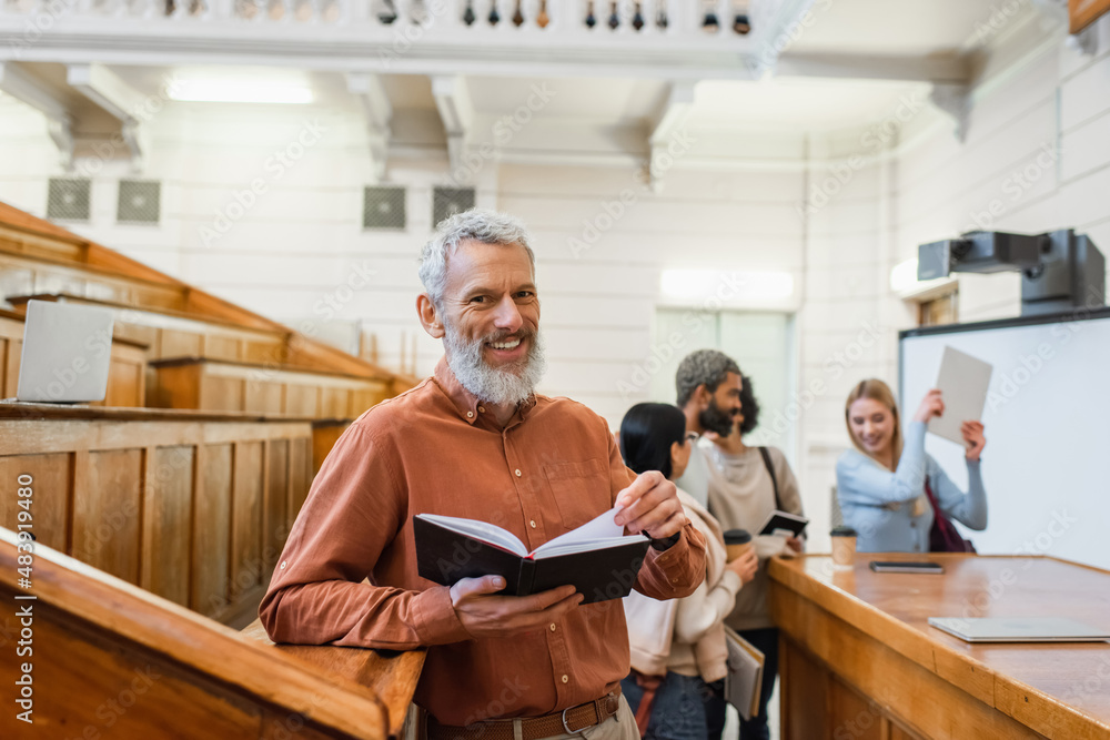 Naklejka premium Positive mature teacher holding notebook near blurred students in university auditorium
