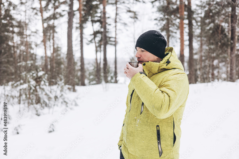 Young man with a cup of tea and thermos in winter snowy forest. White picnic. Drink hot beverage in nature. Local travel. Slow life. Wintertime activity outdoors. Young man with a cup of tea and thermos in winter snowy forest. White picnic. Drink hot beverage in nature. Local travel. Slow life. Wintertime activity outdoors.