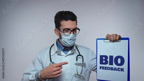 Male doctor wearing eyeglases protective mask and stethoscope showes clipboard with text BIO FEEDBACK over white isilated background photo