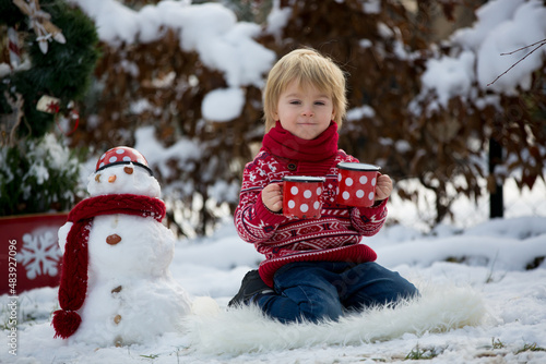 Sweet blond toddler child, boy, playing in garden with snow, making snowman, happy kid winter time