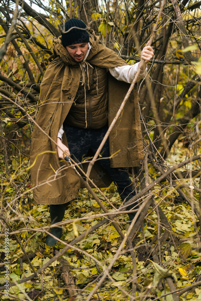 Vertical portrait of serious tourist male wearing raincoat tent chopping small branches with small shovel in forest on overcast cold day. Concept of bushcraft, camping and survival in nature. Vertical portrait of serious tourist male wearing raincoat tent chopping small branches with small shovel in forest on overcast cold day. Concept of bushcraft, camping and survival in nature.