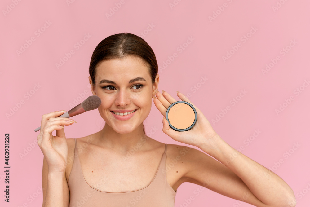 Laughing woman applying powder on her face on a pink background in the studio. Laughing woman applying powder on her face on a pink background in the studio.