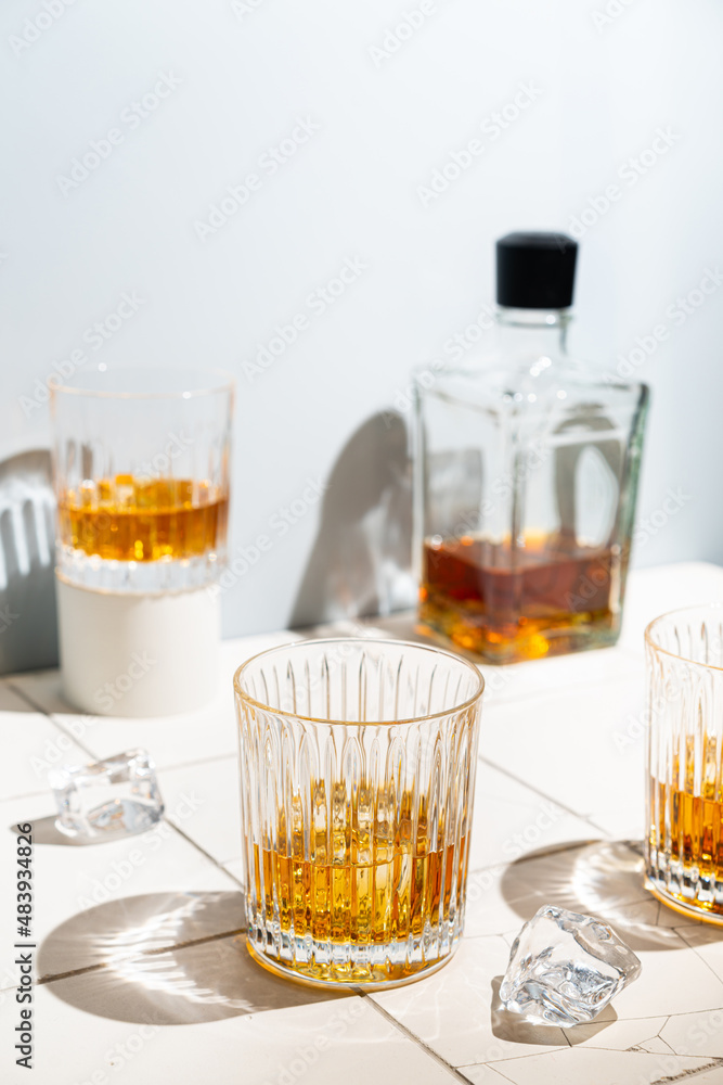 Whiskey with ice in glasses and bottle, white background with hard light, shadows and sun glare, copy space Whiskey with ice in glasses and bottle, white background with hard light, shadows and sun glare, copy space