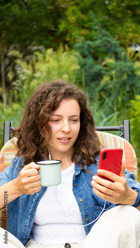 Beautiful young curly woman holding morning coffee cup,looking at mobile phone,sitting on chair at home green backyard garden outdoor.Brunette girl read news,sharing data on social media,chatting,sms
