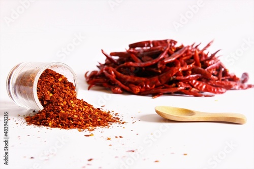 Chili pepper powder cup lying on the floor with wooden spoon and pile of chili peppers beside. stock photo isolate on white front view copy space  