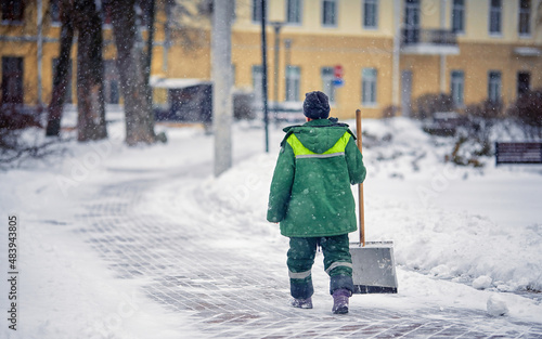 Worker with snow shovel sweep and remove snow, snow removal city service. Janitor in uniform with snowshovel in hands ready to clean snowy sidewalk and walkway during blizzard.