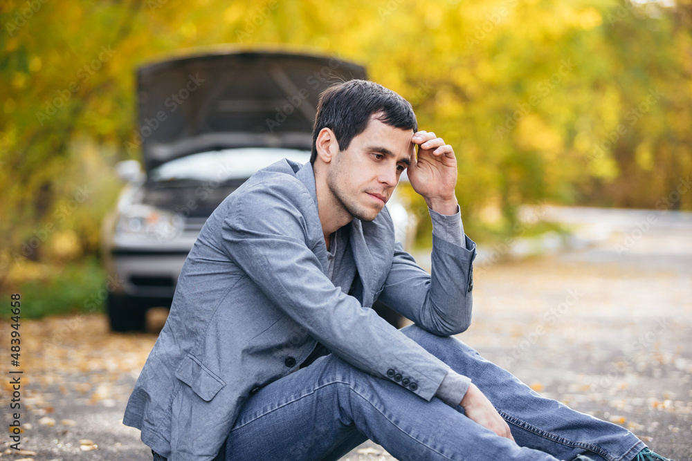 A man in sadness sits on the road in front of a broken car with an open hood A man in sadness sits on the road in front of a broken car with an open hood