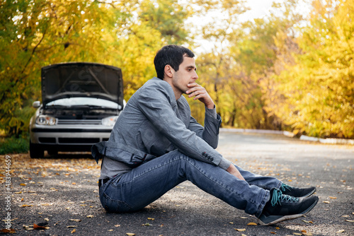 A man in sadness sits on the road in front of a broken car with an open hood photo