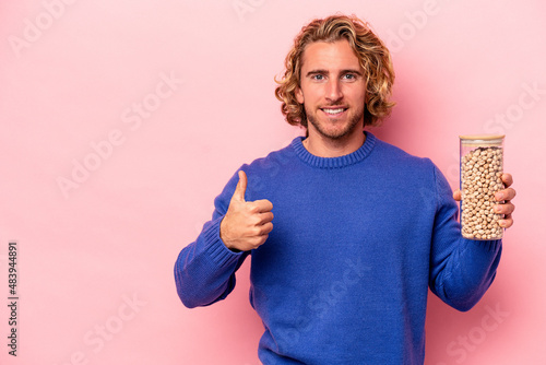 Young caucasian man holding chickpeas isolated on pink background smiling and raising thumb up