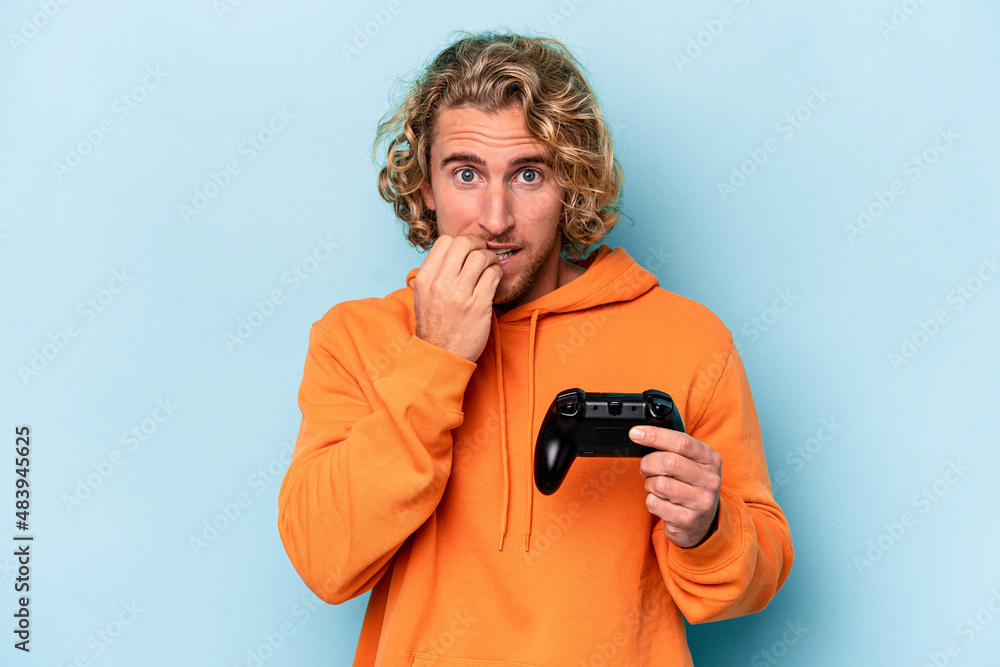 Fototapeta premium Young caucasian man playing with a video game controller isolated on blue background biting fingernails, nervous and very anxious.