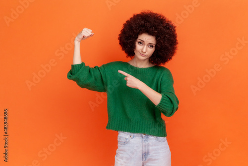 I am strong! Woman with Afro hairstyle wearing green sweater raising hand showing biceps, looking at camera with confidence pride, feeling power. Indoor studio shot isolated on orange background.