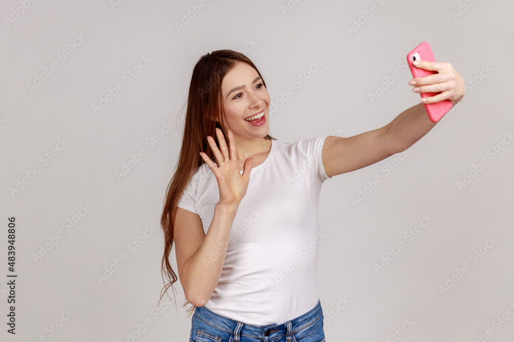 Portrait of happy friendly woman taking selfie and waving hand, gesturing hello, communicating on video call, online chatting, wearing white T-shirt. Indoor studio shot isolated on gray background. Portrait of happy friendly woman taking selfie and waving hand, gesturing hello, communicating on video call, online chatting, wearing white T-shirt. Indoor studio shot isolated on gray background.
