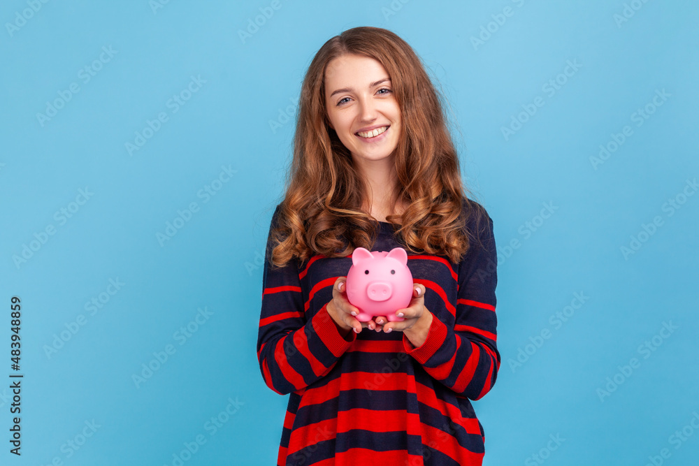 Beautiful woman wearing striped casual style sweater, holding pink piggy bank, looking at camera with charming smile, best variant for saving money. Indoor studio shot isolated on blue background. Beautiful woman wearing striped casual style sweater, holding pink piggy bank, looking at camera with charming smile, best variant for saving money. Indoor studio shot isolated on blue background.