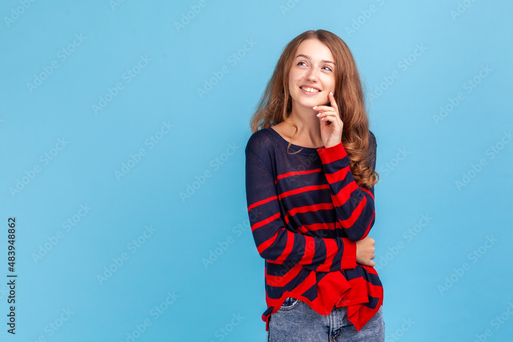 Woman wearing striped casual style sweater standing and looking to the side with thoughtful expression, imagining something pleasant, fantasizing. Indoor studio shot isolated on blue background. Woman wearing striped casual style sweater standing and looking to the side with thoughtful expression, imagining something pleasant, fantasizing. Indoor studio shot isolated on blue background.