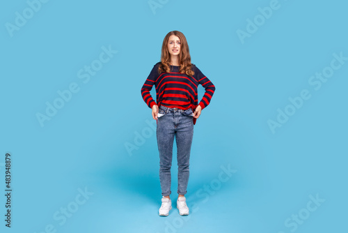 Full length portrait of poor woman, turning out empty pockets, worried about debts, no cash for living, wearing striped casual style sweater. Indoor studio shot isolated on blue background.