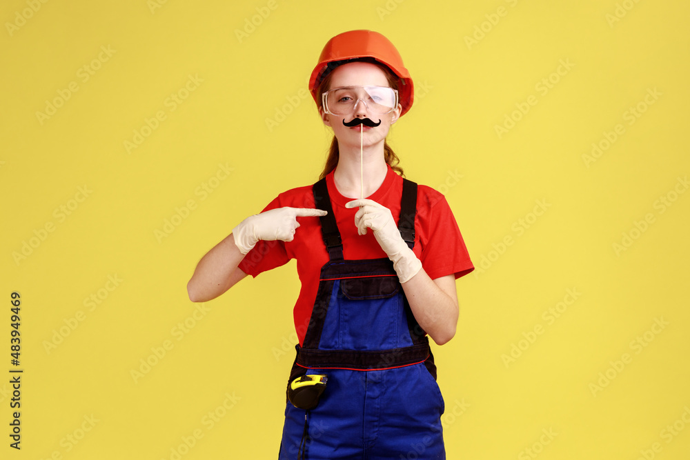 Portrait of funny confident builder woman pointing at mustache on stick, looking at camera, wearing overalls and protective helmet. Indoor studio shot isolated on yellow background.