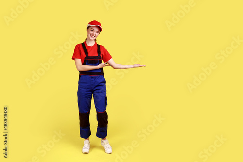 Full length portrait of happy worker woman presenting copy space for advertisement on her palm, looking at camera with smile, wearing overalls and cap. Indoor studio shot isolated on yellow background
