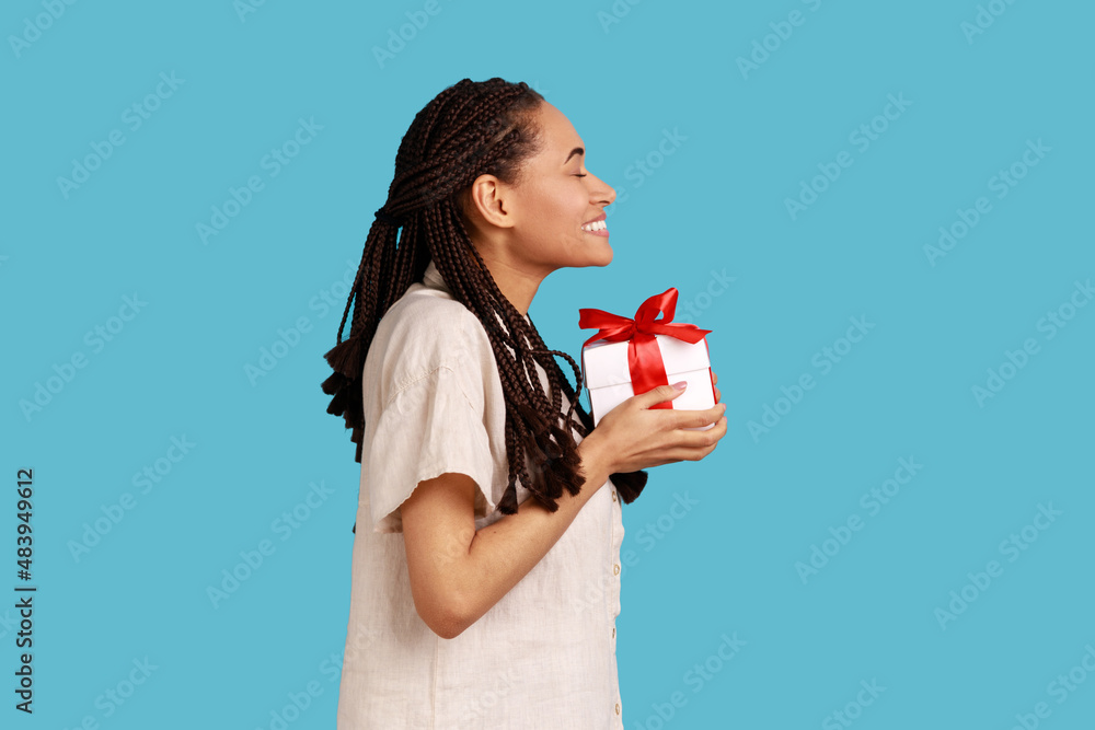 Side view of cute charming woman with black dreadlocks holding present box, congratulating, extremely happy to get gift, wearing white shirt. Indoor studio shot isolated on blue background. Side view of cute charming woman with black dreadlocks holding present box, congratulating, extremely happy to get gift, wearing white shirt. Indoor studio shot isolated on blue background.