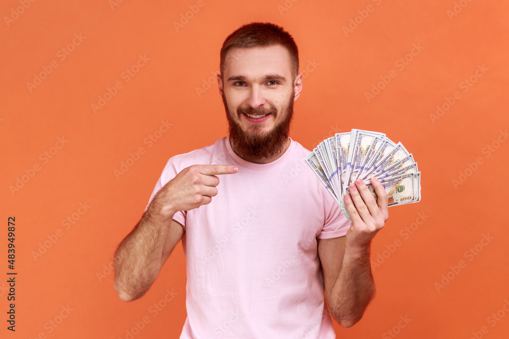 Portrait of bearded man pointing at dollar banknotes and looking with happy expression, financial savings, wearing pink T-shirt. Indoor studio shot isolated on orange background. Portrait of bearded man pointing at dollar banknotes and looking with happy expression, financial savings, wearing pink T-shirt. Indoor studio shot isolated on orange background.