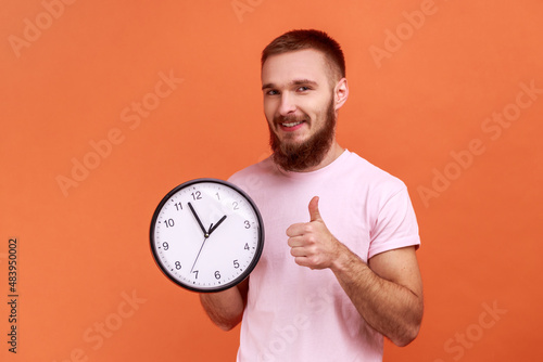 Portrait of bearded man showing thumbs up holding big wall clock, looking at camera with toothy smile, time management, wearing pink T-shirt. Indoor studio shot isolated on orange background.