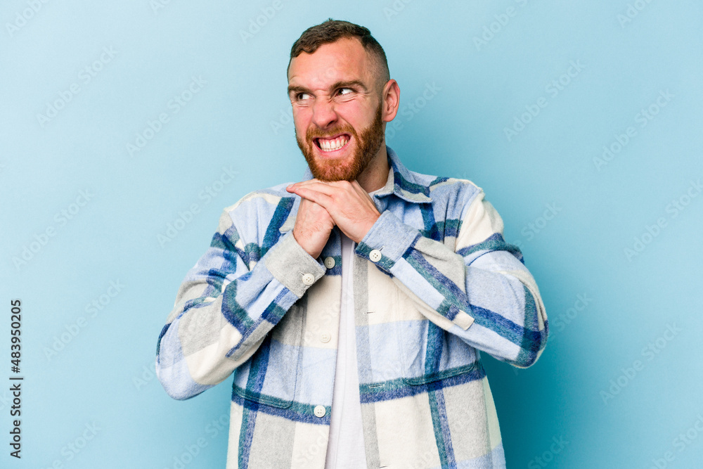 Young caucasian man isolated on blue background keeps hands under chin, is looking happily aside. Young caucasian man isolated on blue background keeps hands under chin, is looking happily aside.