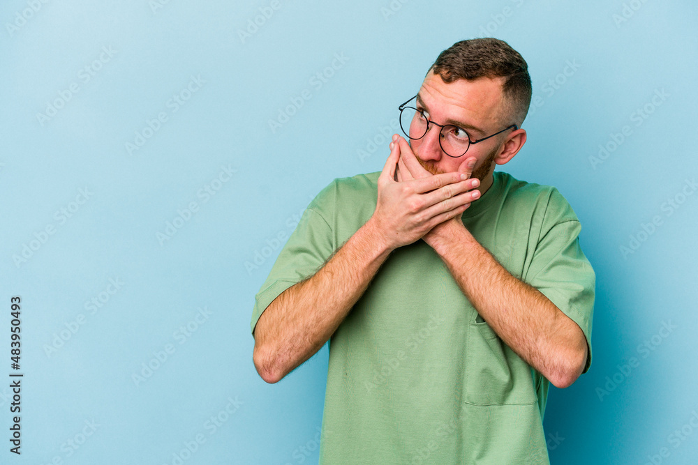 Young caucasian man isolated on blue background thoughtful looking to a copy space covering mouth with hand. Young caucasian man isolated on blue background thoughtful looking to a copy space covering mouth with hand.