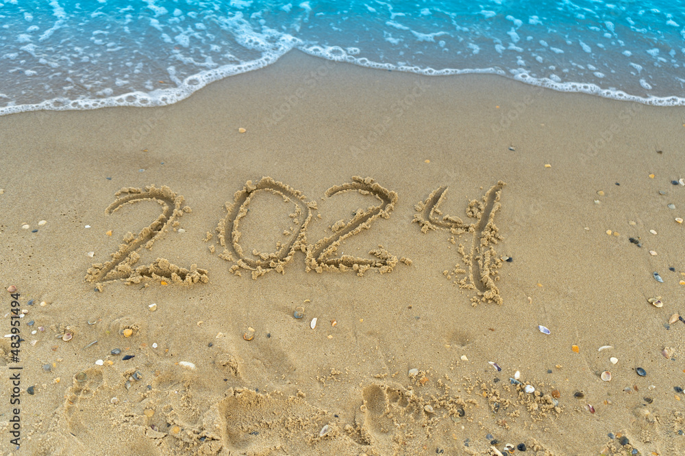 The numbers 2024 are written on a sandy seashell beach. Wave and surf