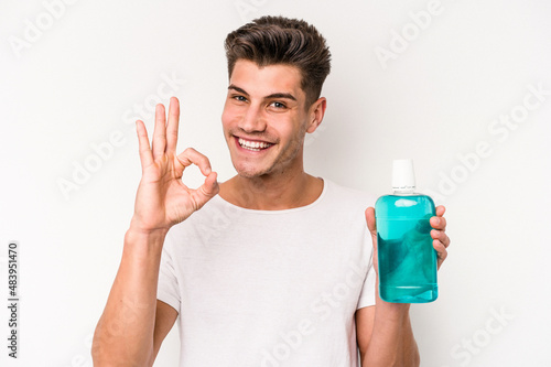 Young caucasian man holding mouthwash isolated on white background cheerful and confident showing ok gesture.