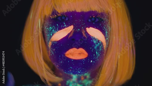 Neon body art. Close up of young woman wearing wig smearing orange paint on her cheeks, dancing in ultraviolet lights