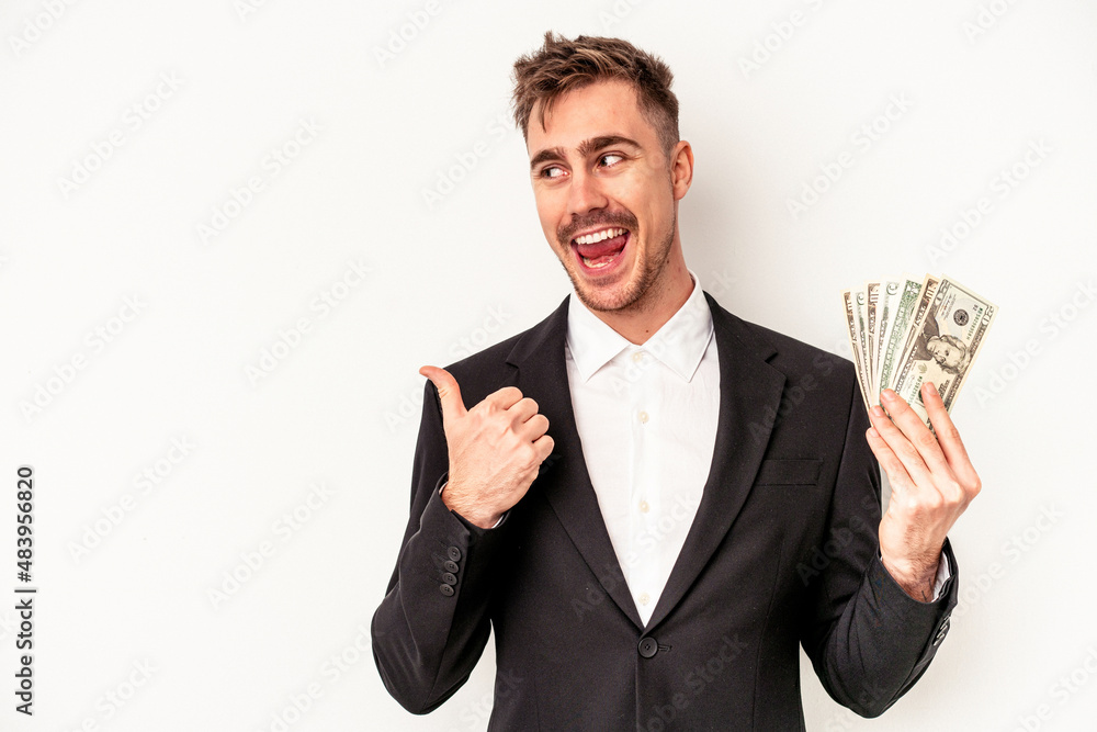 Young caucasian business man holding bank notes isolated on white background points with thumb finger away, laughing and carefree. Young caucasian business man holding bank notes isolated on white background points with thumb finger away, laughing and carefree.