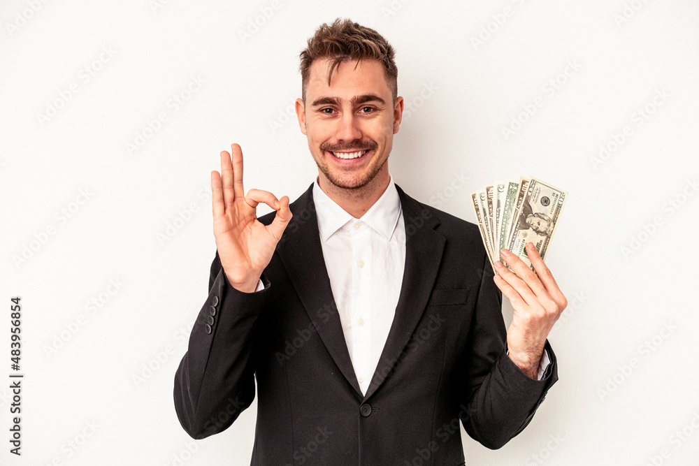 Young caucasian business man holding bank notes isolated on white background cheerful and confident showing ok gesture. Young caucasian business man holding bank notes isolated on white background cheerful and confident showing ok gesture.