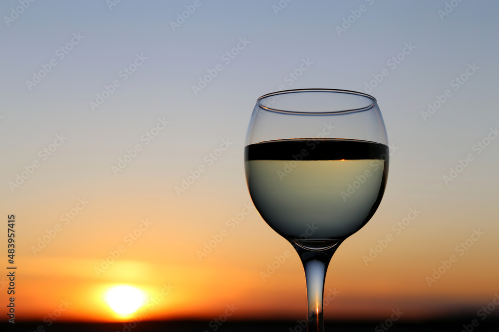 Glass with white wine on sunset background, evening sun and sky reflected in the glass. Concept of celebration, wine industry