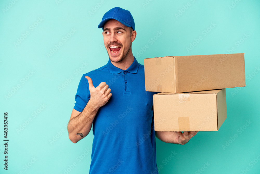 Young delivery caucasian man isolated on blue background points with thumb finger away, laughing and carefree. Young delivery caucasian man isolated on blue background points with thumb finger away, laughing and carefree.