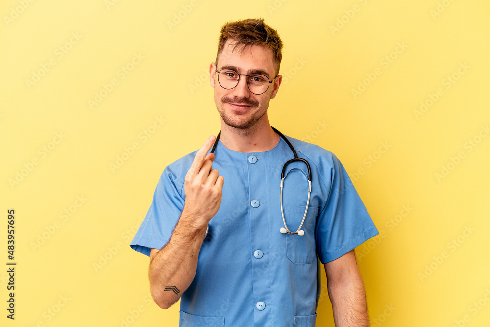 Young nurse caucasian man isolated on yellow background pointing with finger at you as if inviting come closer. Young nurse caucasian man isolated on yellow background pointing with finger at you as if inviting come closer.