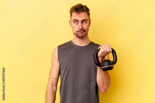 Young sport man holding a dumbbell isolated on yellow background confused, feels doubtful and unsure.