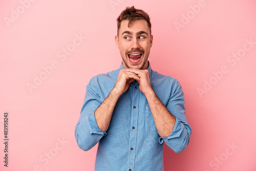 Young caucasian man isolated on pink background keeps hands under chin, is looking happily aside.