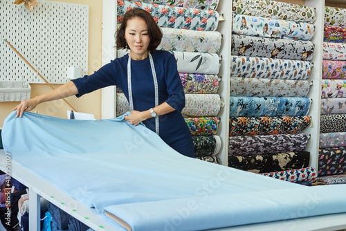 Beautiful Asian woman dressmaker unrolls a roll of blue fabric for cutting. Small business concept. © evafesenuk