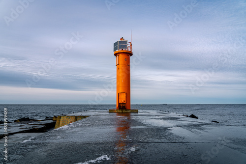 An orange lighthouse at the end of the Mangalsala or Eastern pier in Riga, Latvia where the river Daugava flows into the Baltic Sea. Winter storm, cold weather, ice and snow.